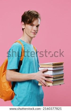 vertical photo of a student in a blue t-shirt with a stack of new books in his hands, standing on a pink background