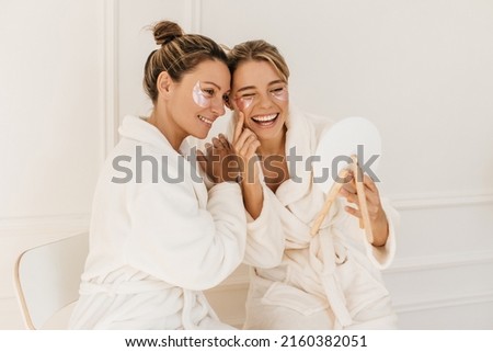 Pretty caucasian young women wear bathrobes admire themselves looking in mirror on white background. Blondes use eye patches for dark circles. Beauty and personal care, skin and face care concept