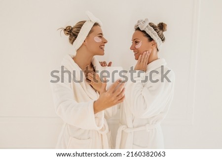 Young blonde caucasian girls laughing looking at each other holding phone on white background. Women wear robes, hairbands, and patches under their eyes. Beauty needs care concept Royalty-Free Stock Photo #2160382063