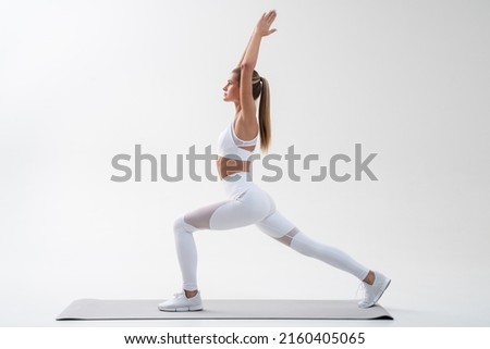 Full body side view picture of attractive young female doing yoga or pilates standing in warrior pose isolated on white background, training on mat in comfortable leggings and sport top