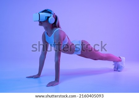 Full body picture in neon light of athletic young female in sportswear wearing vr goggles and headphone standing in plank position, doing workout exercises remotely. Sports and high cyber technology