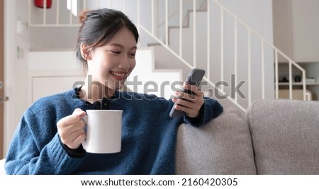 Portrait of a smiling asian women sitting at sofa and drink coffee looking at smartphone.
