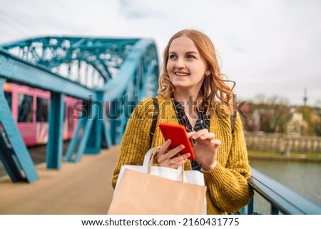Portrait of cheerful attractive young caucasian woman with blonde hair in dress smiling in camera with teeth, holding shopping bags and smartphone in hands, catting with friend.