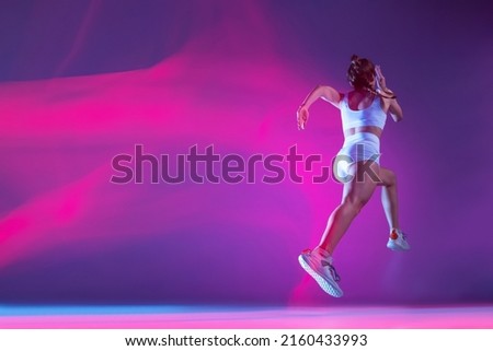 back view. Professional female athlete, runner training isolated on blue studio background in mixed pink neon light. Sportive girl in white sportswear practicing in run or jogging.