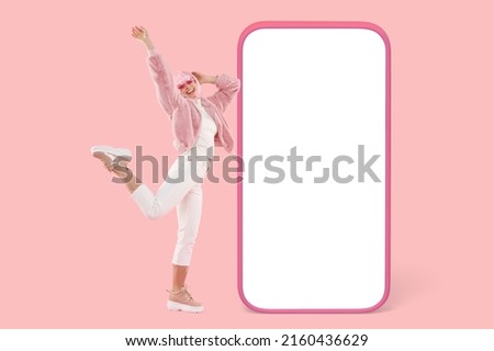 Full length picture of free young woman in wig and pinkish short coat flying in jump throwing hands up over pink background in front of huge phone mock up with copy space for your advertising content Royalty-Free Stock Photo #2160436629