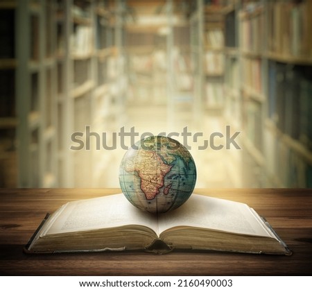 Old globe lying on an open book against the background of bookshelves in a library. Selective focus. Retro style. Science, education, travel, vintage background. Education history and geography team. Royalty-Free Stock Photo #2160490003