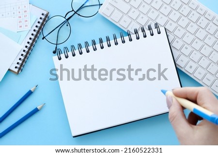 Notepad, white keyboard, pen, woman's hands writing in empty notebook at the blue desk. Flat lay top view.