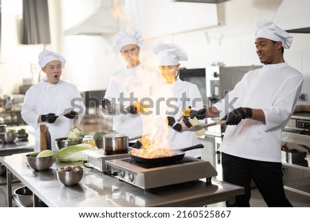 Multiracial team of professional cooks in uniform preparing meals for a restaurant in the kitchen. Latin guy burning pan, european cooks making sauce and asian chef managing the process. Teamwork and Royalty-Free Stock Photo #2160525867
