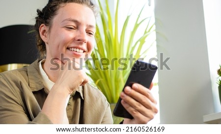 Close up overjoyed young caucasian woman using phone, browsing apps, sitting indoors near window and green plant. Reading good news in message, chatting or shopping online, having fun with gadget