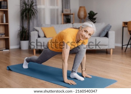Athletic aged woman stretching her legs, doing runner's lunge yoga pose on home workout, exercising in living room. Domestic training during covid lockdown