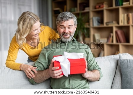 Loving wife surprising her middle aged husband with gift at home, greeting him with birthday or anniversary, happy woman giving present box to excited man, celebrating valentine's day together