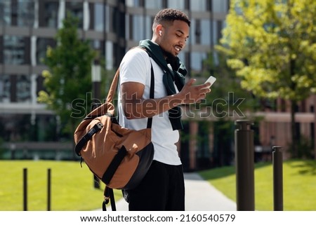Stylish Young Black Man In Wireless Headphones Using Cell Phone Walking Down The Street In The Urban City. Cheerful Trendy Guy Listening To Favorite Music Spending Time Outdoors, Walking Resting Alone Royalty-Free Stock Photo #2160540579