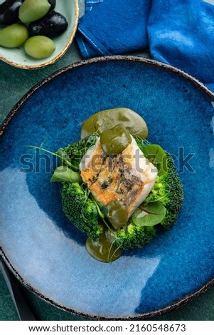 Fried cod fillet with broccoli, herbs and basil sauce on a blue plate, top view