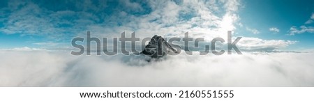 Panoramic aerial photograph taken with a drone. The peak of Beriáin, in the Andía mountain range, rises among the white clouds. In the background a deep blue sky and the sun rising through the clouds. Royalty-Free Stock Photo #2160551555