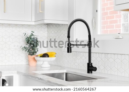 A farmhouse kitchen sink detail shot with a black faucet, mosaic tile backsplash, marble countertops, and white cabinets. Royalty-Free Stock Photo #2160564267