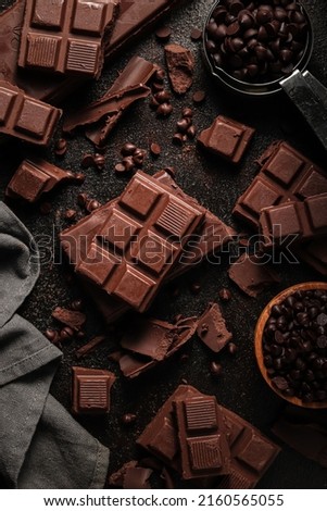 chocolate bars, milk chocolate slices and chocochips on a wooden bowl and a measuring cup. on a dark wooden background with copy space, top view angle (flatlay), blurry background 