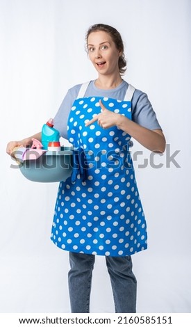 european housewife in a blue kitchen apron, jeans and a t-shirt on a white background, full length portrait. a woman holds a basin with detergents, smiles, points at us or at the basin