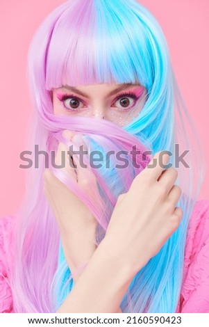 Hairstyling, hair dye. Portrait of a beautiful girl with bright pink makeup hiding her face with her colored purple-blue hair and looking with surprise at the camera. Pink background. Beauty concept.