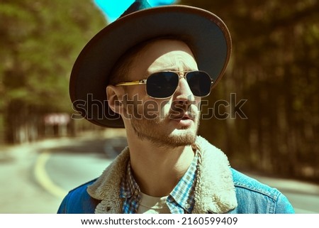 Portrait of a handsome bearded man in denim jacket, a hat and black sunglasses standing by the highway. Denim fashion. Road adventures, hitchhiking, autotrip.  Royalty-Free Stock Photo #2160599409