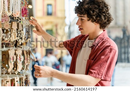 Curly young man looking at the souvenirs at the market. Typical souvenir shop selling souvenirs and handicrafts at the market. Souvenirs at the neck and jewellery  Royalty-Free Stock Photo #2160614197