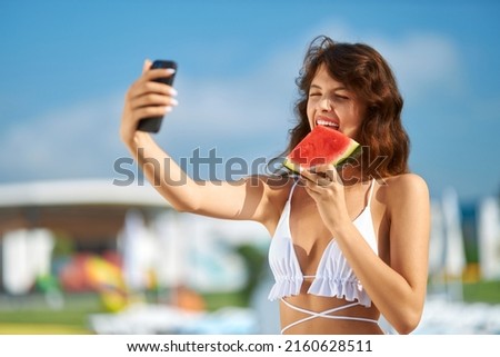 Pretty lady in bikini taking selfie, while biting tasty slice of watermelon on vacation. Portrait view of cute girl eating watermelon, holding sell phone to take funny photo at day. Concept of selfie.