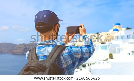 Back view of young tourist man backpack  in  using smartphone take picture at View of blue church dome in Oia village,Santorini,Greece