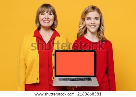 Happy woman 50s in red shirt have fun with teenager girl 12-13 years old. Grandmother granddaughter hold use work on laptop pc computer blank screen workspace area isolated on plain yellow background.