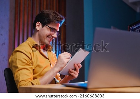 Young smiling professional fun content creator IT specialist programmer man in shirt work at office on laptop pc computer using tablet search information browse internet. Program development concept Royalty-Free Stock Photo #2160668603