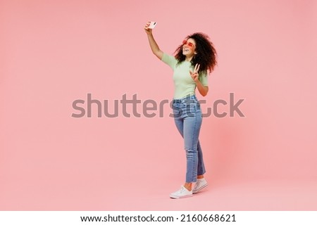 Full size happy fancy young curly latin woman 20s wears casual clothes do selfie shot on mobile cell phone post photo on social network isolated on plain pastel light pink background studio portrait