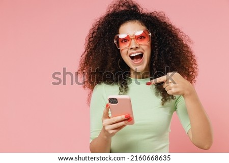 Jubilant happy young curly latin woman 20s wears mint t-shirt sunglasses hold in hand use mobile cell phone pointing forefinger on screen isolated on plain pastel light pink background studio portrait Royalty-Free Stock Photo #2160668635