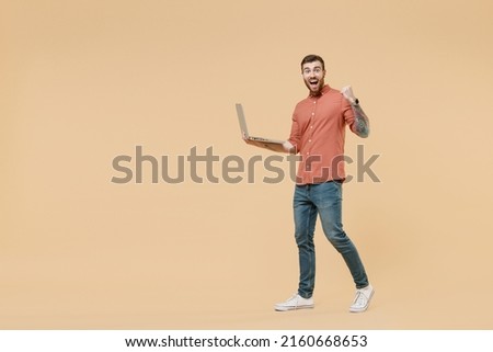 Full size body length fun tattooed young brunet man 20s short haircut wears brown shirt go stride hold use laptop pc computer clench fists say yes isolated on pastel orange background studio portrait