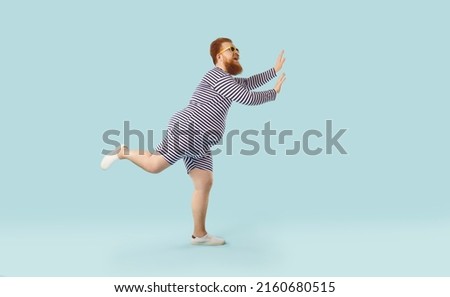 Profile view of funny fat ginger man wearing striped swimsuit and sunglasses dancing and having fun isolated on light blue colour background. Summer holiday, vacation, beach party concept Royalty-Free Stock Photo #2160680515
