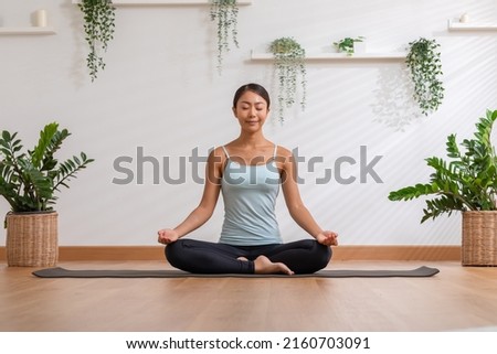 Calm of wellness Asian young woman sit on yoga mat doing breathing with yoga lotus pose.Yoga meditation of young healthy woman relax and comfortable at white cozy home,Yoga Exercise Wellness Concept