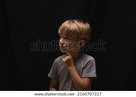 Profile portrait of pensive handsome blonde little boy in gray T-shirt with finger near mouth on black background. Mute boy speaks with gestures. Counts in mind or remembers poem. Kid think or compos. Royalty-Free Stock Photo #2160707227