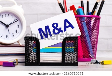 The office desk has diaries, an alarm clock, stationery, and a white card with the text M AND A. Business concept.