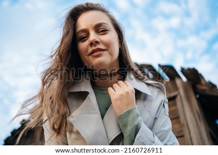 beautiful girl with long hair in a grey trench coat next to an old wooden house blue sky background outdoors in spring
