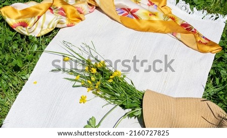 A straw hat, a bouquet of wild flowers, a silk handkerchief lie on a rug on the green grass. The concept of outdoor recreation. Bright sunlight, selective focus, blurry background.