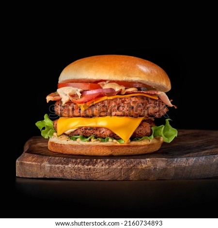 Big sandwich - hamburger burger with beef, tomato, pickled cucumber and fried bacon. Craft beef burger on wooden tray isolated on black background.