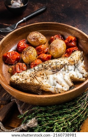 Baked halibut fish with roasted tomato and potato in wooden plate. Dark background. Top view.