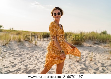 stylish attractive slim smiling woman on beach in summer style fashion trend outfit carefree and happy, feeling freedom, wearing yellow printed dress boho style chic and sunglasses Royalty-Free Stock Photo #2160751197