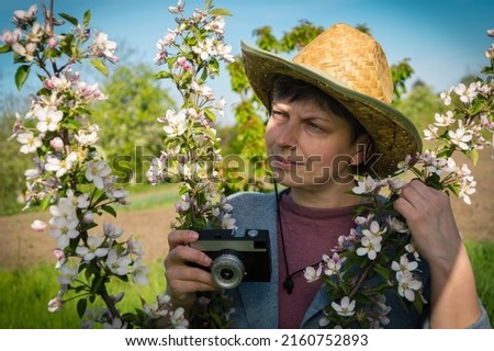 Middle-aged woman in a straw hat with a vintage camera photographing a blossoming branch of an apple tree in the garden.