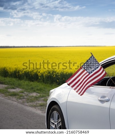 American flag is hanging out of window of car against backdrop of flowering yellow rapeseed field. United States Independence Day July 4th. Pride, freedom, patriotism, democracy. Travel around country Royalty-Free Stock Photo #2160758469