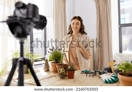 blogging, gardening and housework concept - happy woman or blogger with camera and pot flowers recording tutorial video and showing thumbs up at home