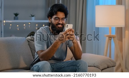 Smiling Arabian Indian man bearded male looking in smartphone smile texting with friend girlfriend messaging chatting at home sitting on couch at night playing game in mobile app watching funny video Royalty-Free Stock Photo #2160770193