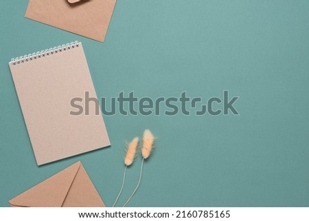 Home office desk. Brown blank notebook, craft envelopes and dried lagurus grass. Pastel turquoise background. Top view, flat lay. Women's workspace.