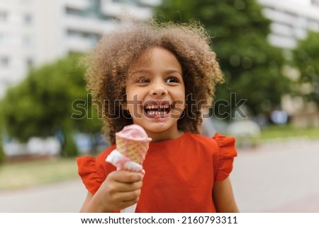 A black laughing child in a red dress eats ice cream on a hot day. Ice cream in a waffle cone. A happy and contented child at summer. Royalty-Free Stock Photo #2160793311