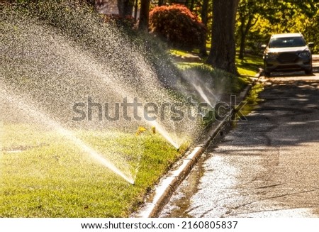 Sprinklers running in residential neighborhood with shady street with car parked in background - selective focus Royalty-Free Stock Photo #2160805837