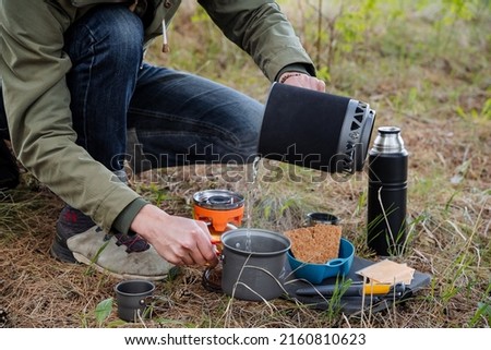A tourist on a hike cooks food on a gas burner, camping in the forest, pour water into a pot for boiling tea. Camping equipment, tourist breakfast, outdoor lunch, High quality photo