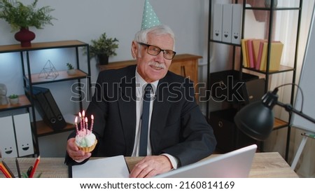 Happy senior businessman celebrating lonely birthday in office, holding small cake with candles, making a wish. Elderly success grandfather man wearing festive cap celebrate anniversary party alone