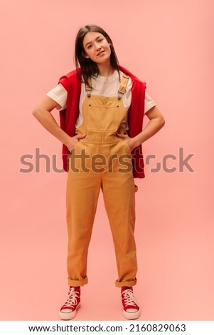 Full-length calm young caucasian brunette girl in casual clothes against pink background. Teenager looks into camera and keeps hands in jumpsuit pockets. Lifestyle, feminine beauty concept